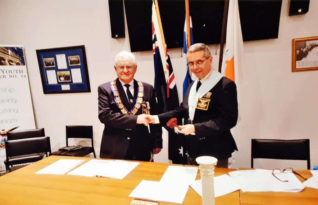 Formal ceremony of AHEPA Victoria featuring two members shaking hands, one presenting an award or plaque, in a setting with Australian flags.